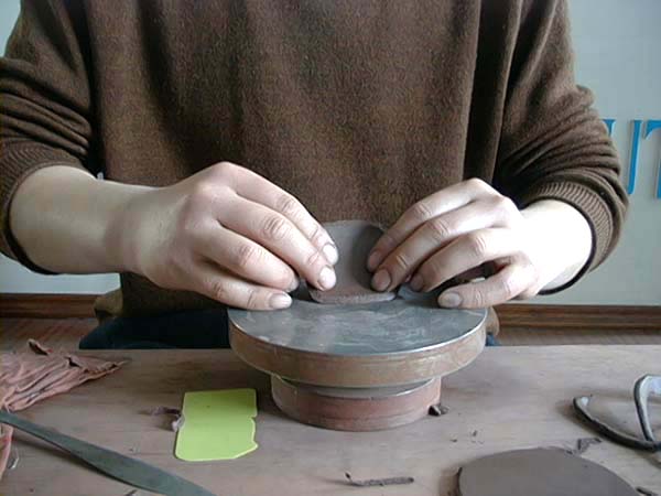 30、以打好之泥片，套上制作设计好的纸样，切割出壶盖上部的泥片大小及形状，然后放上转盘，双手并用地，弯曲出方形壶盖之曲线弧度。